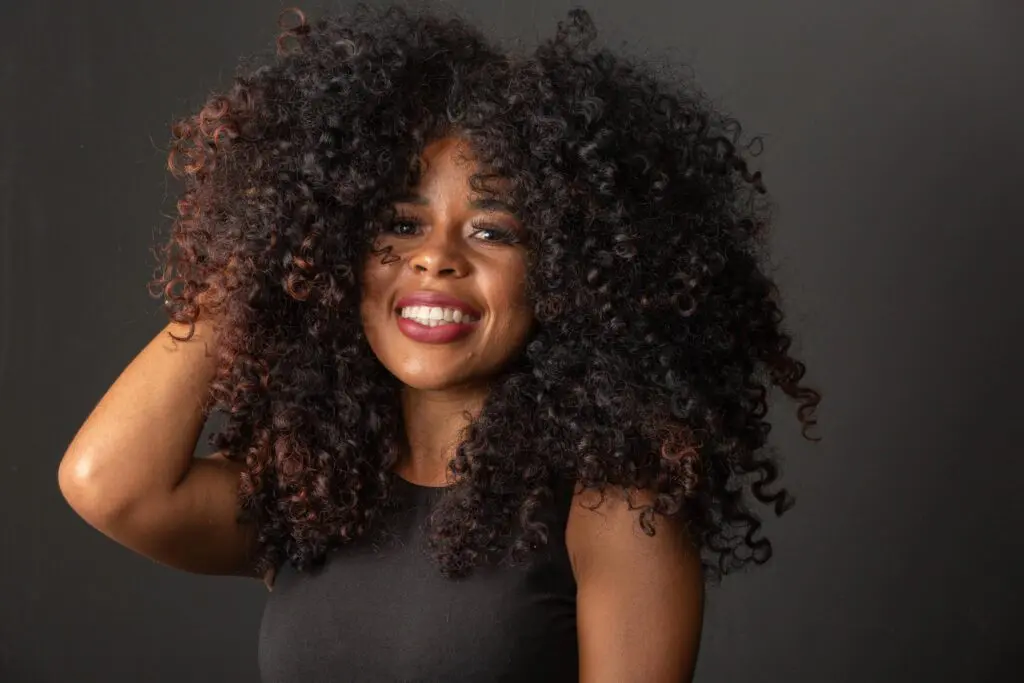 curly.hair A woman with voluminous curls smiles brightly, wearing a black sleeveless top and posing against a plain dark background. Her hand is lifted to her hair, embodying the confidence of a curly hair stylist.