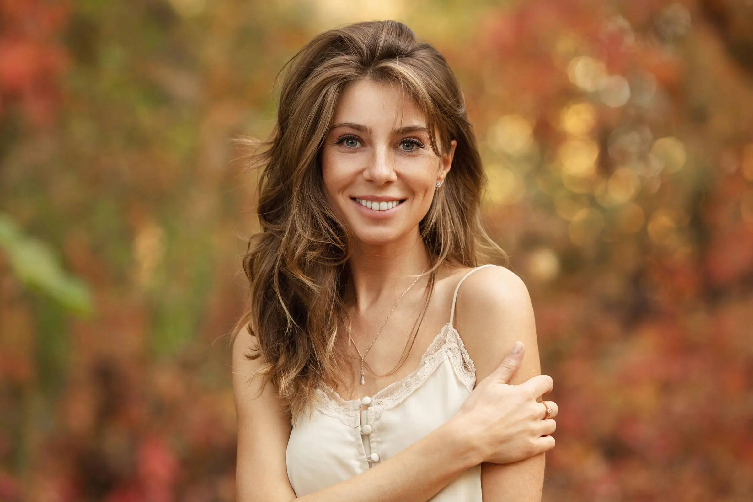livedin A young woman with long brown hair and a light camisole smiles at the camera, standing outdoors in front of a blurred background with lived in color from vibrant autumn foliage.