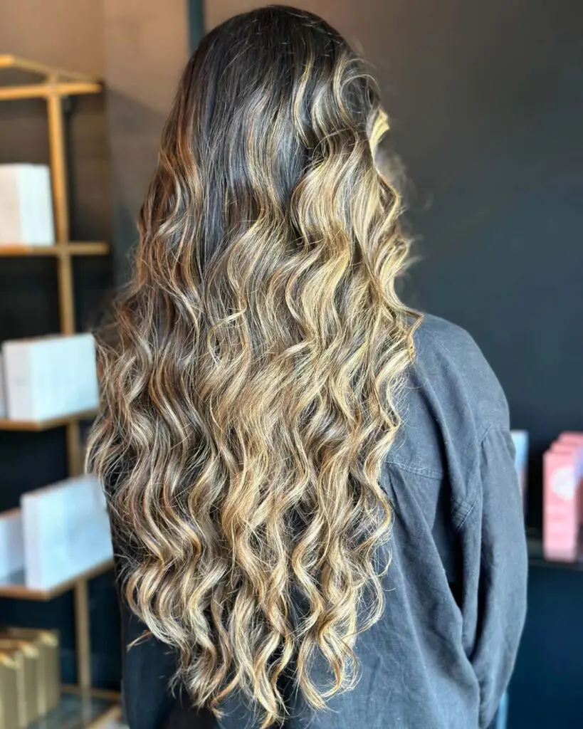 A client showcasing ultra-long hair styled in a voluminous, wavy blowout with high-contrast golden blonde dimensional highlights, beautifully crafted at Sunday Salon in Raleigh.