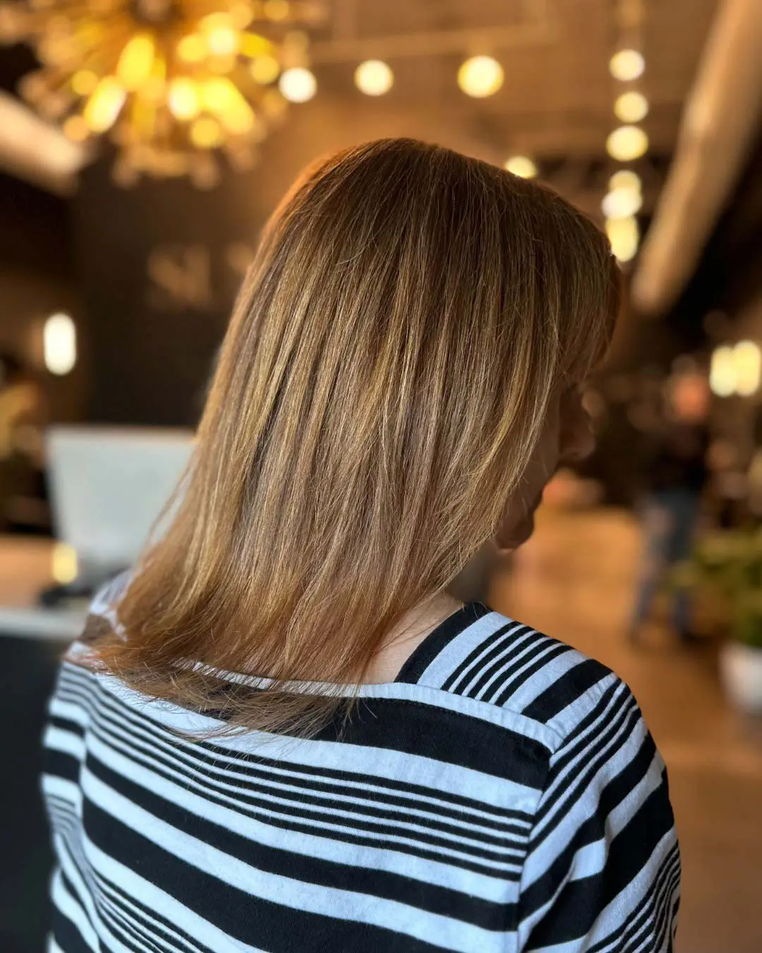 A client showcasing shoulder-length hair styled in a sleek and smooth blowout, featuring dimensional warm honey blonde highlights, created at Sunday Salon in Raleigh.