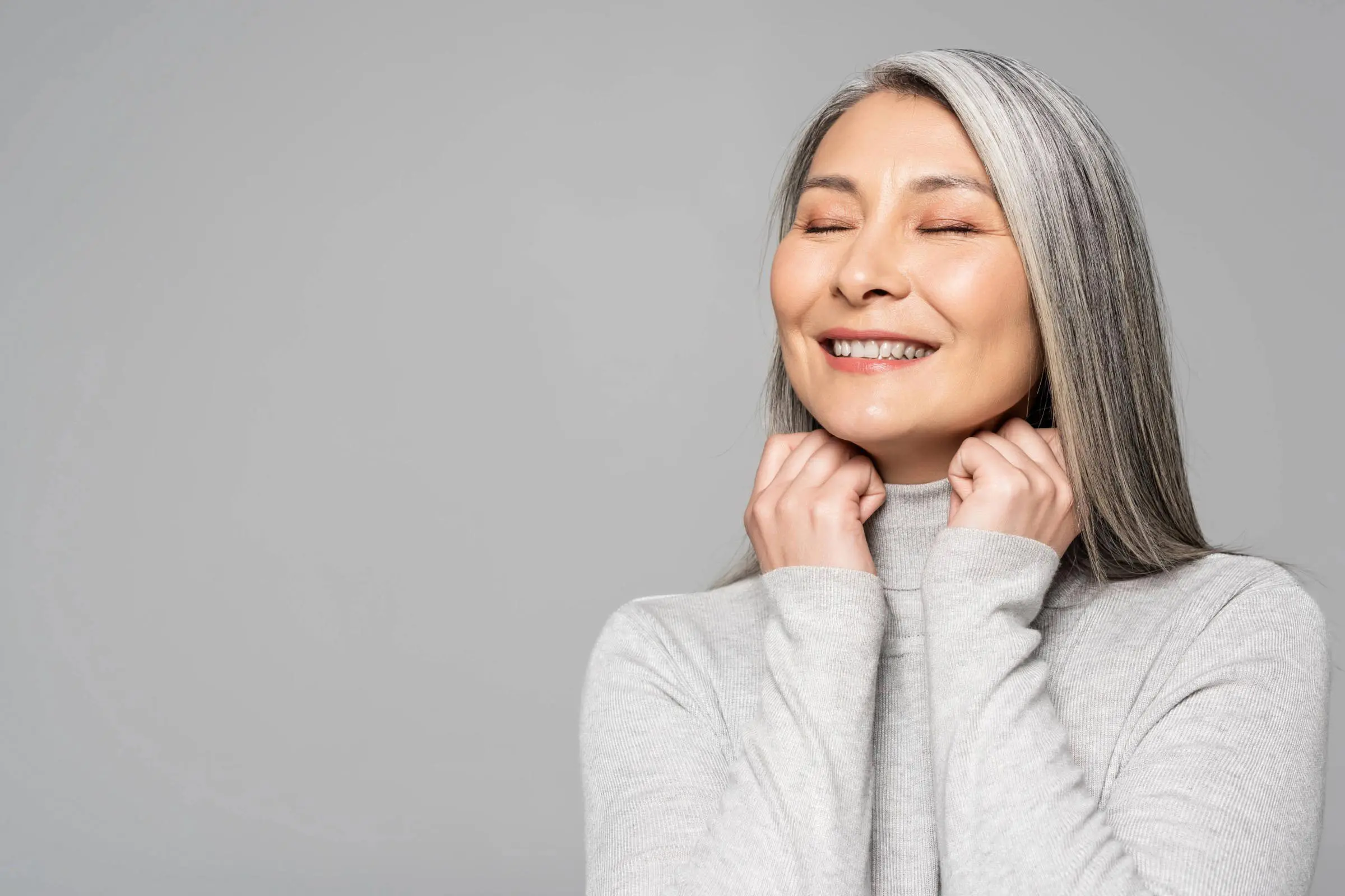 A woman with long gray hair smiles with her eyes closed, gently holding her hands near her chin. She is wearing a light gray turtleneck sweater and stands against a plain gray background, embodying confidence in embracing gray hair solutions.