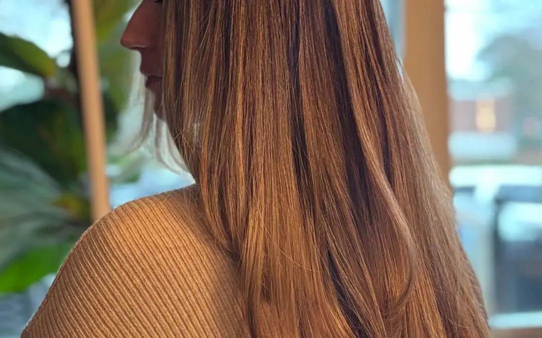 A side profile of a client showing off ultra-long, thick, warm caramel brunette hair styled in a sleek blowout with softly sweeping layered ends at Sunday Salon in Raleigh.