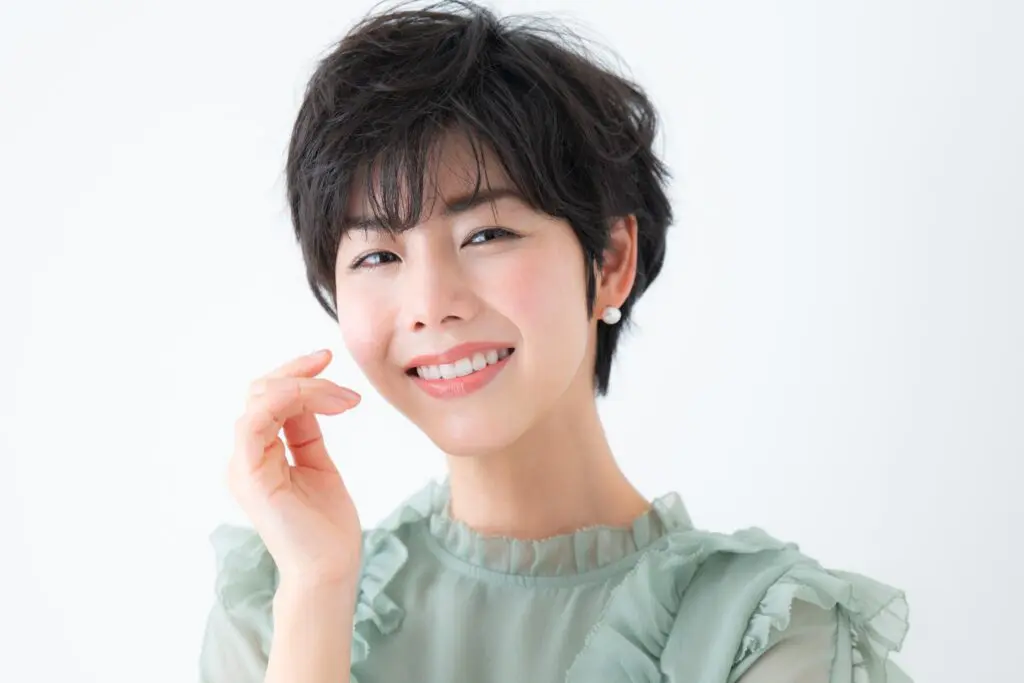 A woman with short, dark hair and pearl earrings smiles warmly at the camera. She is wearing a light green, ruffled top and poses with her right hand gently raised near her face against a white background.