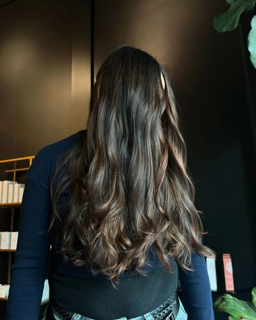 Ultra-Long Dark Brunette with Warm Caramel Balayage A client with ultra-long dark brunette hair featuring dimensional warm caramel balayage highlights, styled in loose waves at Sunday Salon in Raleigh.