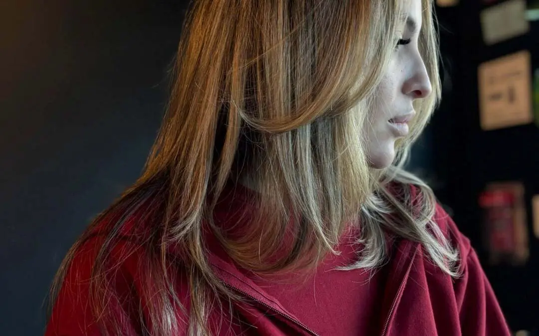 A side profile of a client with mid-length warm honey blonde highlighted hair, styled in a smooth blowout with sweeping face-framing layers at Sunday Salon in Raleigh.