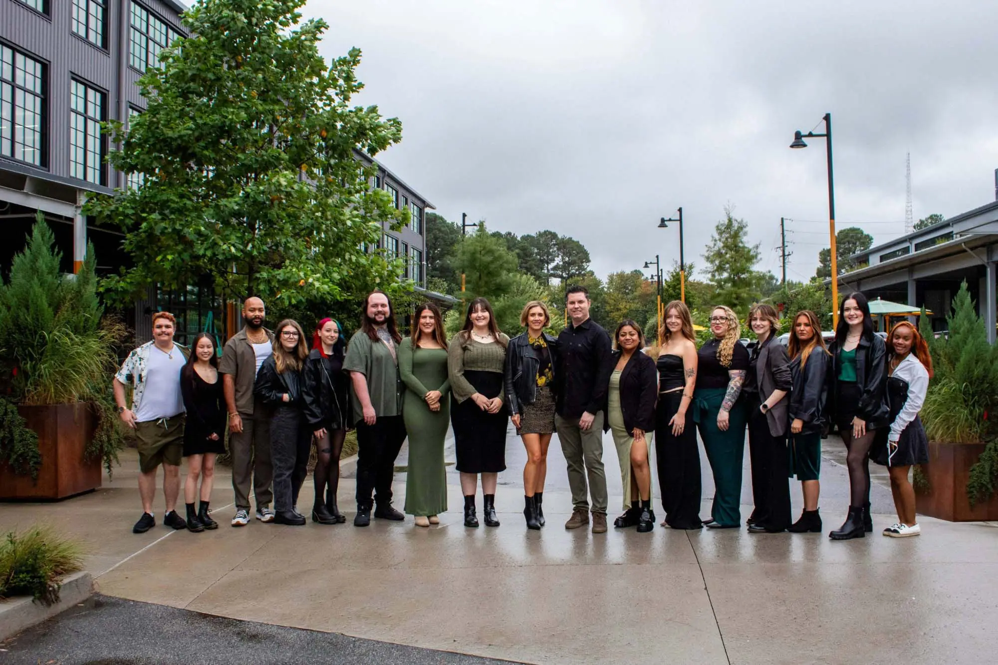 A group of seventeen people stands outdoors on a wet sidewalk, posing for a photo. They are dressed in semi-formal attire, with greenery and modern buildings in the background on a cloudy day.