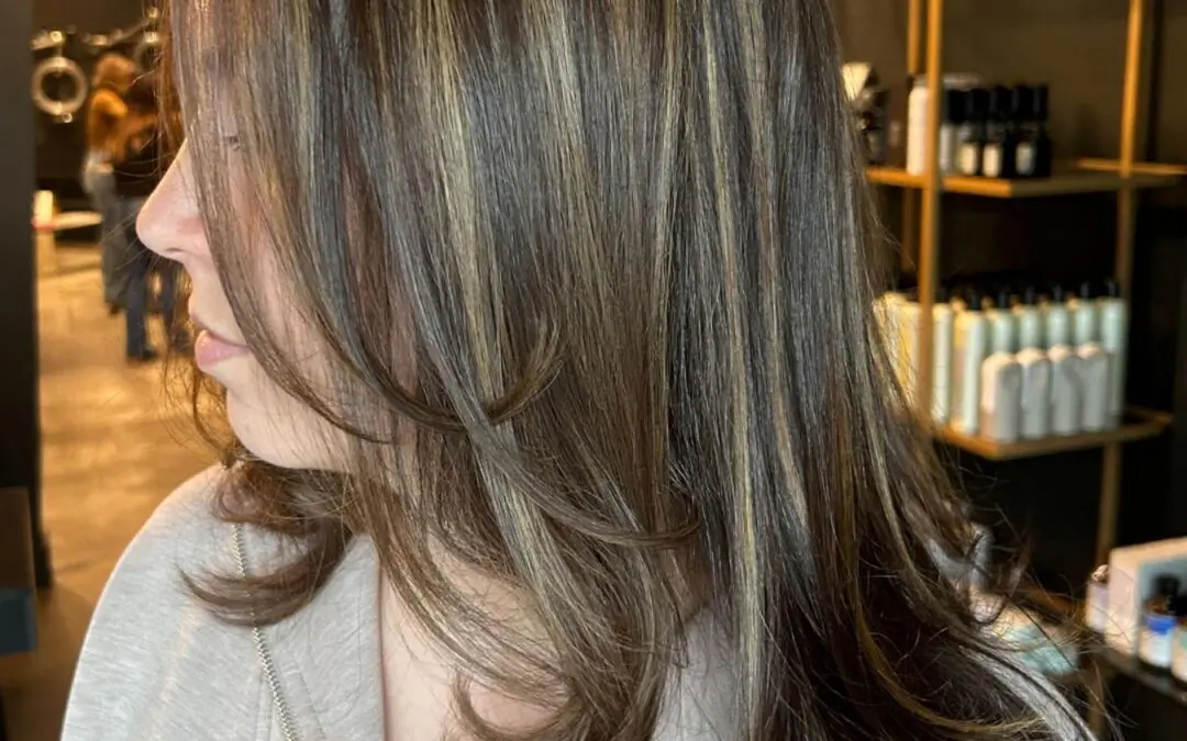 A side profile of a client showcasing long dark brunette hair with dimensional golden blonde highlights, styled in a sleek blowout with flipped face-framing layers at Sunday Salon in Raleigh.