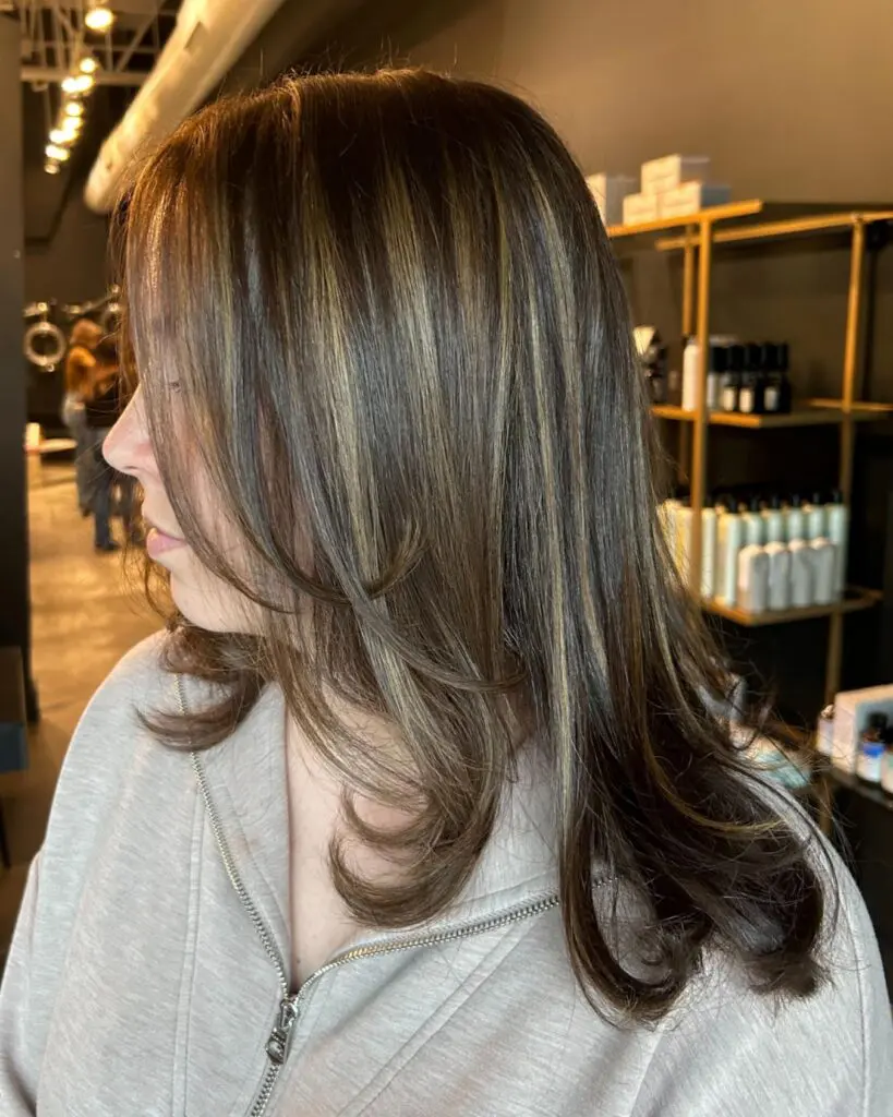Sleek Brunette with Face-Framing Layers and Golden Ribbon Highlights A side profile of a client showcasing long dark brunette hair with dimensional golden blonde highlights, styled in a sleek blowout with flipped face-framing layers at Sunday Salon in Raleigh.
