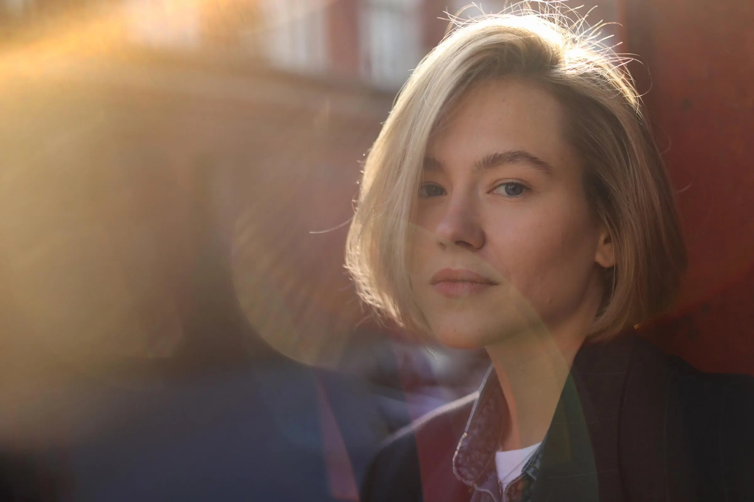 A person with short blonde hair and subtle blonde highlights stands outdoors in soft sunlight, with lens flare partially obscuring the scene and a blurred background, a slight smile on their face.