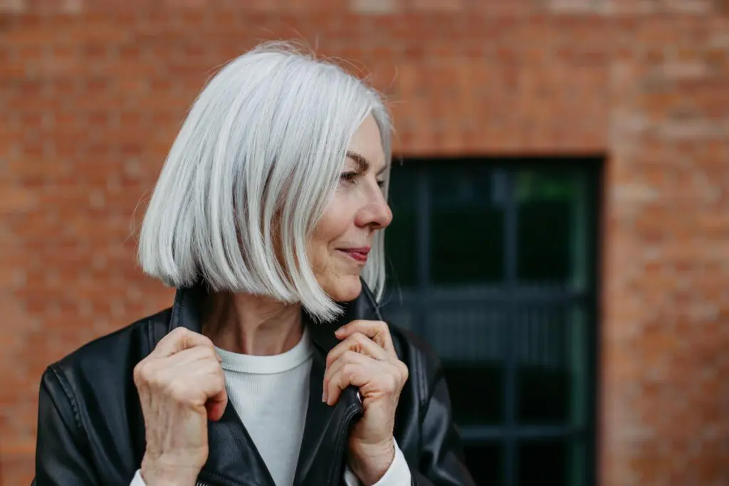 An older woman with chin-length white hair stands outside in front of a brick wall and window, holding the collar of her black leather jacket and looking thoughtfully to the side, embodying confidence and embracing modern gray hair solutions.