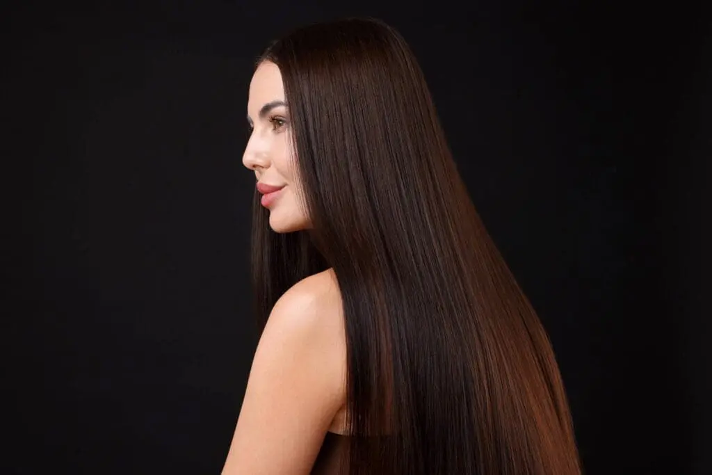 A woman with long, straight, shiny brown hair poses in profile against a black background, highlighting the smooth texture and healthy appearance achieved through keratin treatments.
