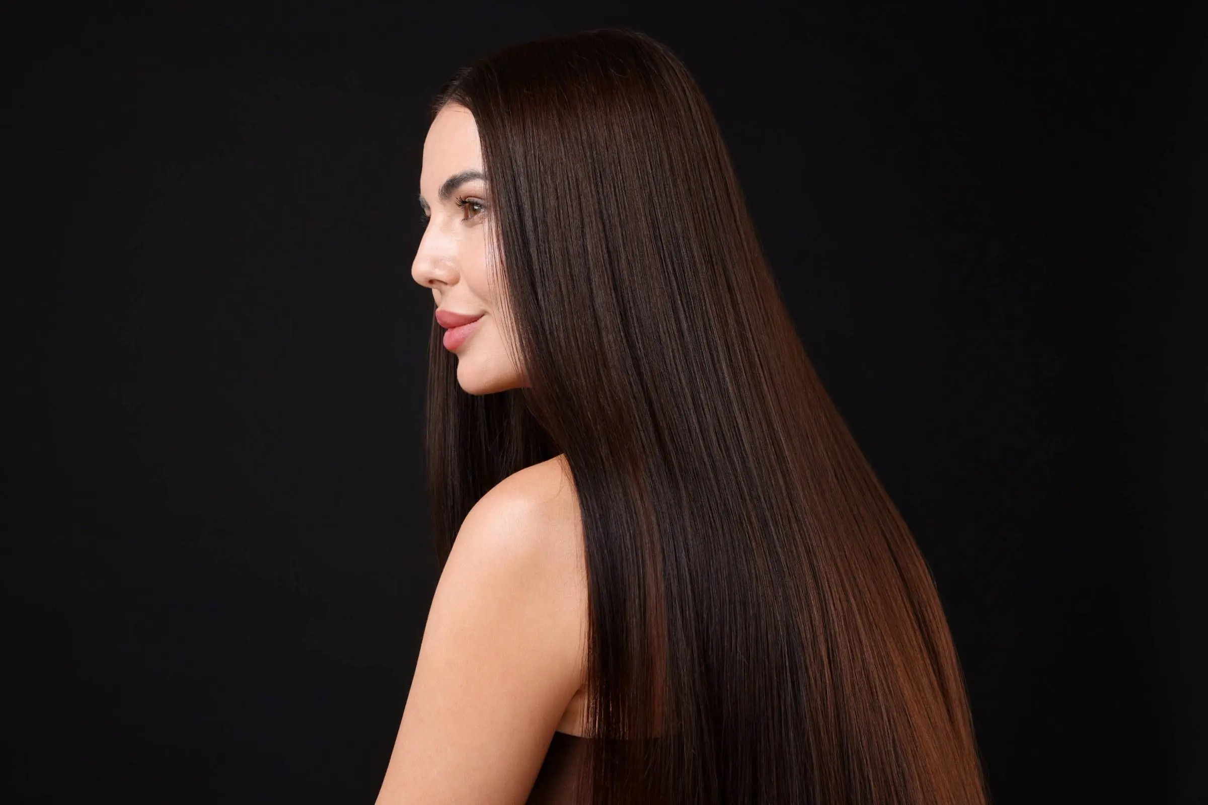 A woman with long, straight, shiny brown hair poses in profile against a black background, highlighting the smooth texture and healthy appearance achieved through keratin treatments.