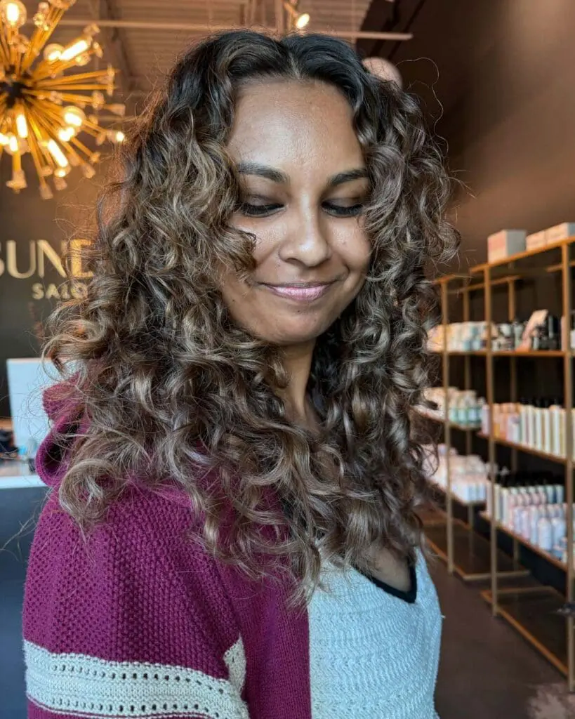 A happy client showcasing long, voluminous, naturally curly hair with defined ringlets. The style features a dark brunette shadow root that beautifully melts into dimensional caramel and light brown highlights, crafted at Sunday Salon in Raleigh.