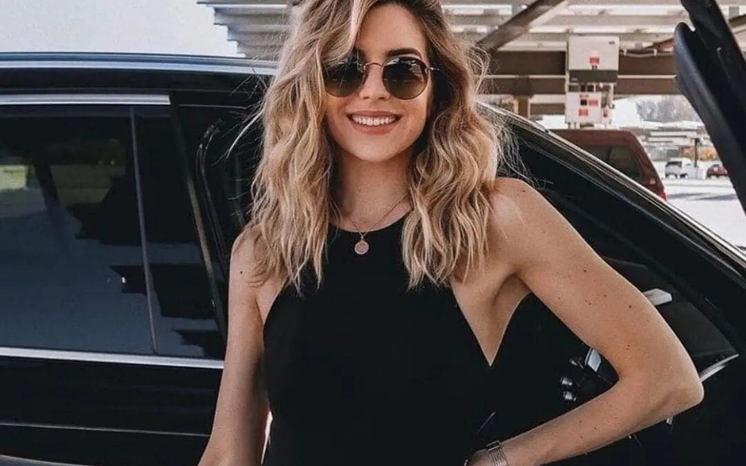 A woman with wavy blonde hair, wearing sunglasses, a black sleeveless top, and black jeans, smiles while standing by an open car door in a sunlit parking area.