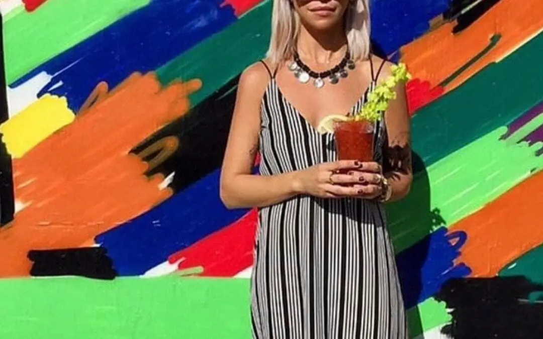A woman standing in front of a colorful mural showcasing a sleek, shoulder-length icy platinum blonde lob with a dark root shadow by Lisa Edwards at Sunday Salon in Raleigh, NC.