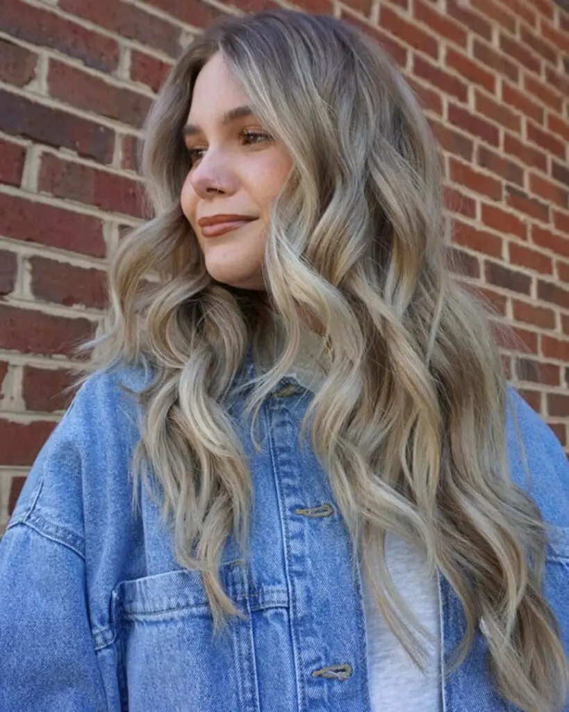 A smiling woman outdoors against a brick wall, showcasing ultra-long wavy hair with a dimensional ash and mushroom blonde balayage and a seamless shadow root at Sunday Salon in Raleigh, NC.