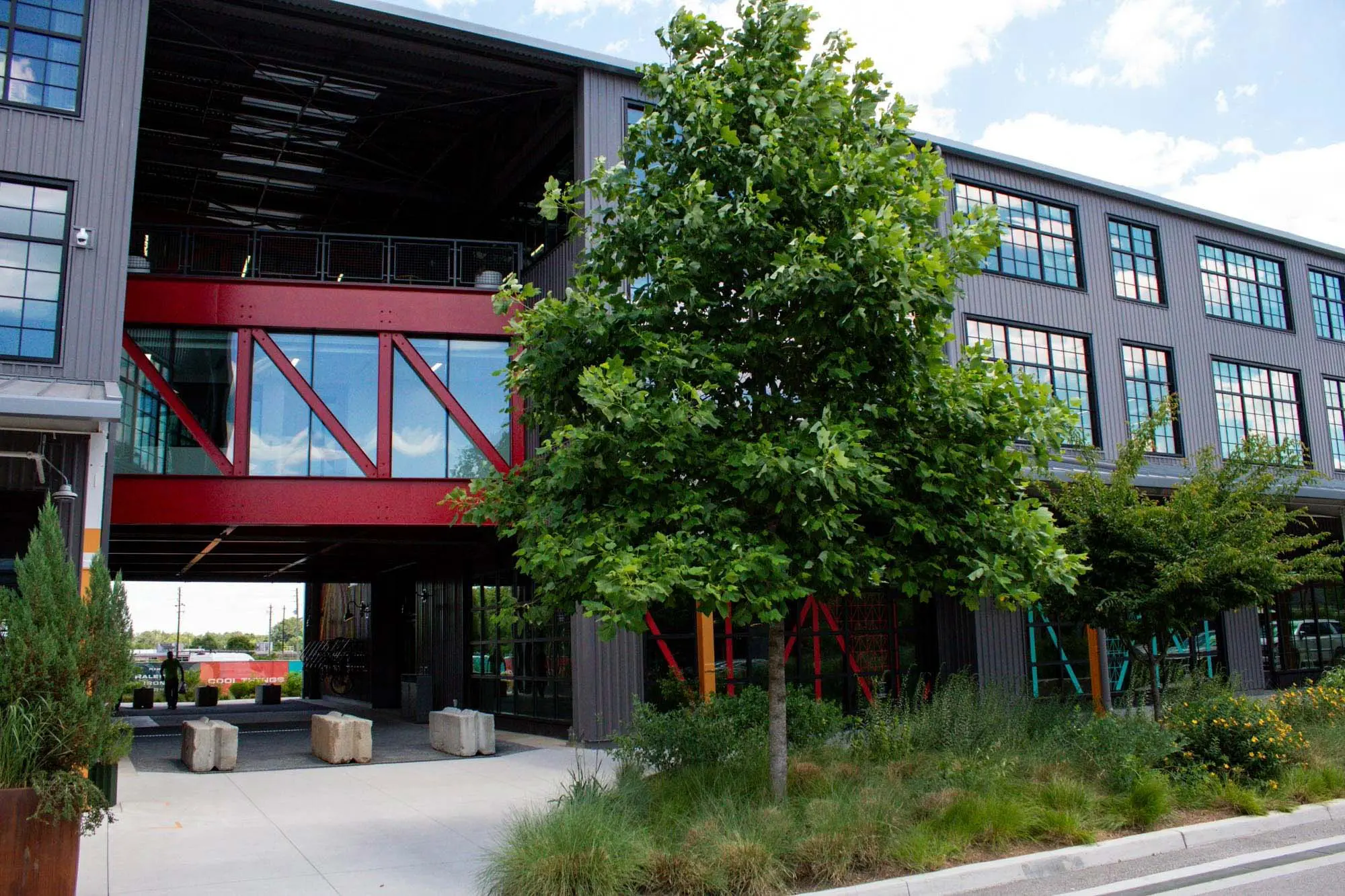 A modern industrial building with large windows, gray exterior, and a red skybridge, surrounded by green trees and landscaped plants along a sidewalk.