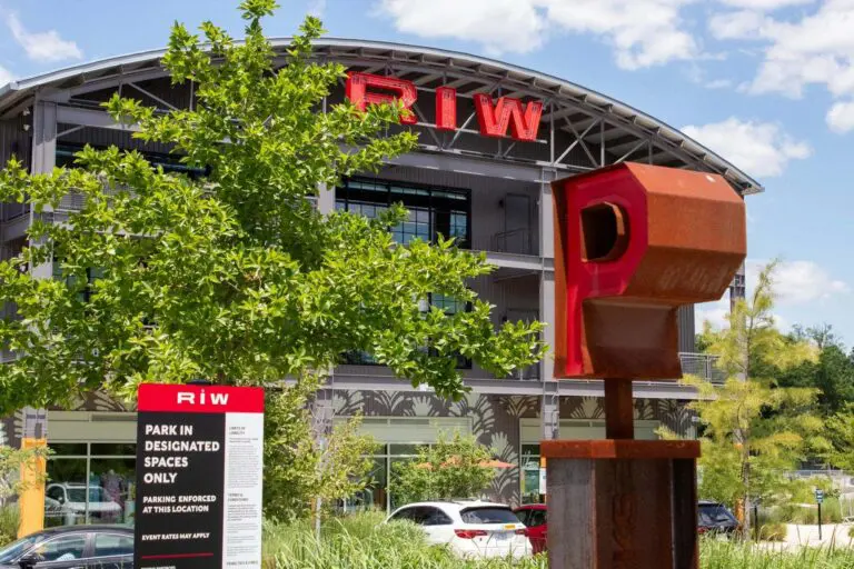 A modern building with a large red “RIW” sign on the roof, a metal sculpture in the foreground, trees, and several parked cars. A white and red parking sign is also visible near the sculpture.