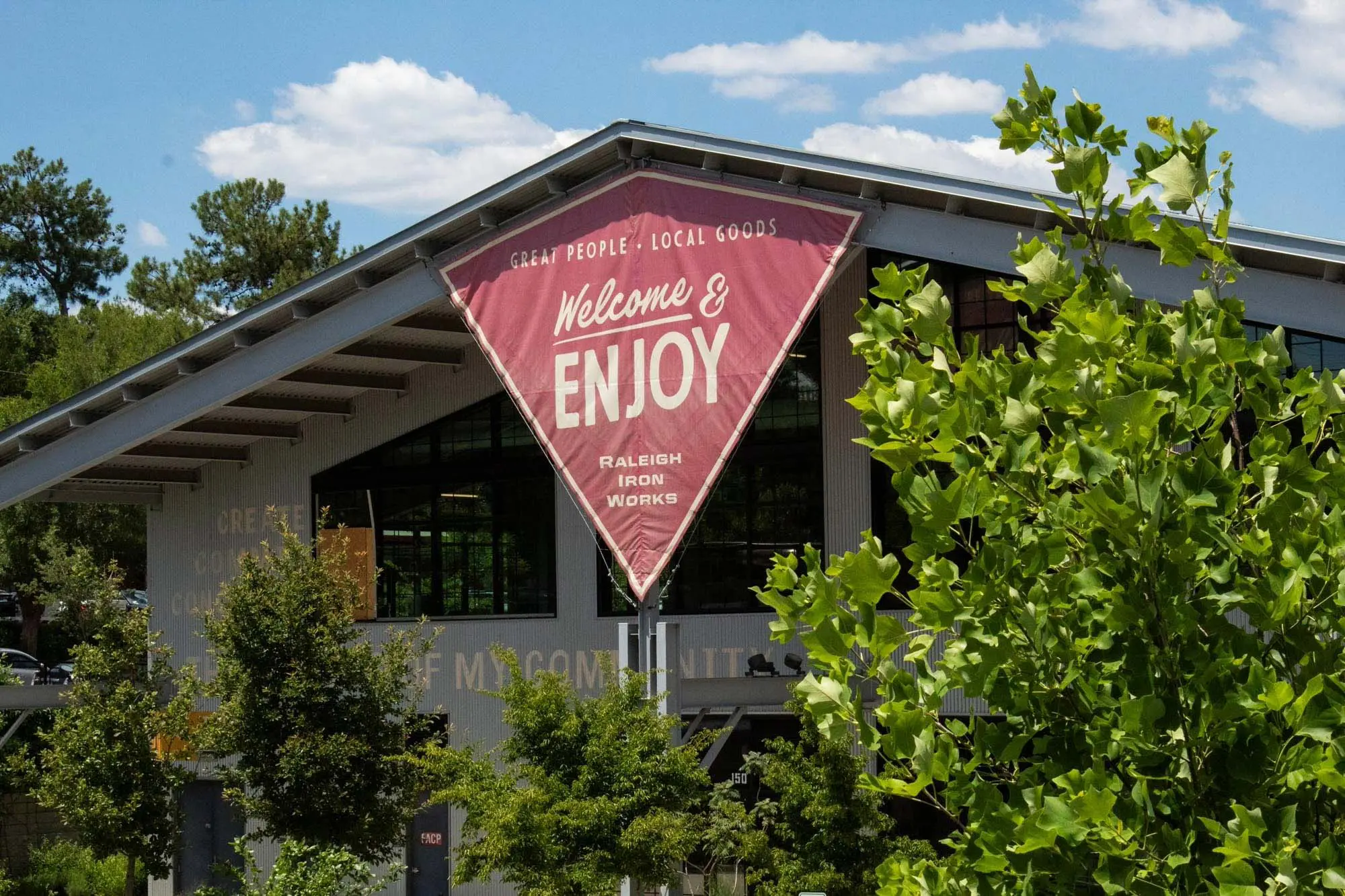 A large red triangular sign reading "Welcome & Enjoy" hangs on the exterior of a modern building surrounded by green trees under a blue sky. The building is labeled "Raleigh Iron Works.