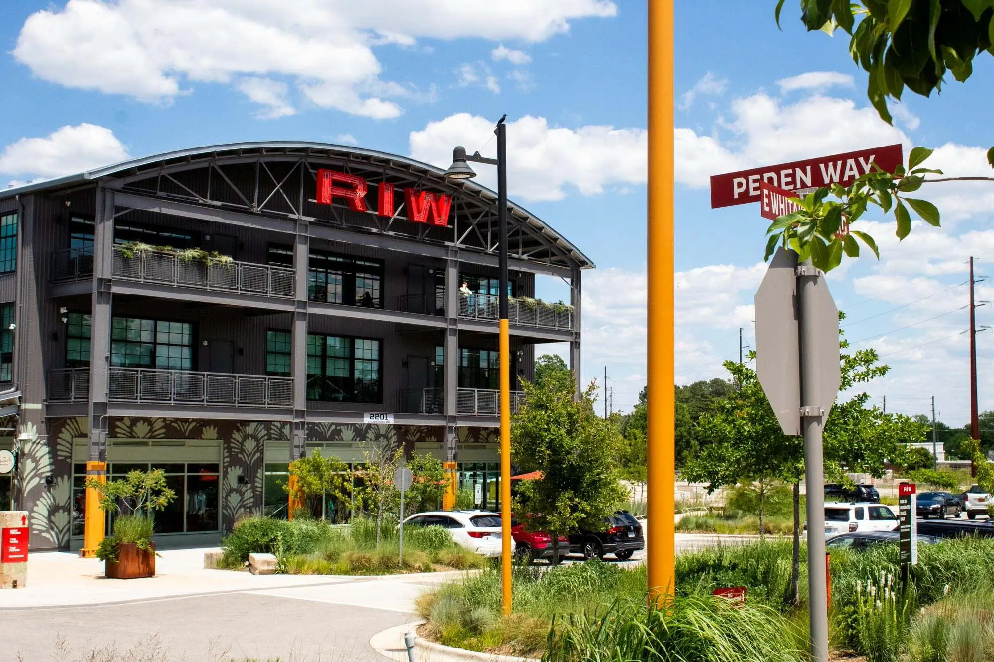 A modern industrial-style building with "RW" in red letters on top, greenery on balconies, and a street sign reading "Peden Way" in the foreground, under a partly cloudy sky. Cars are parked along the street.