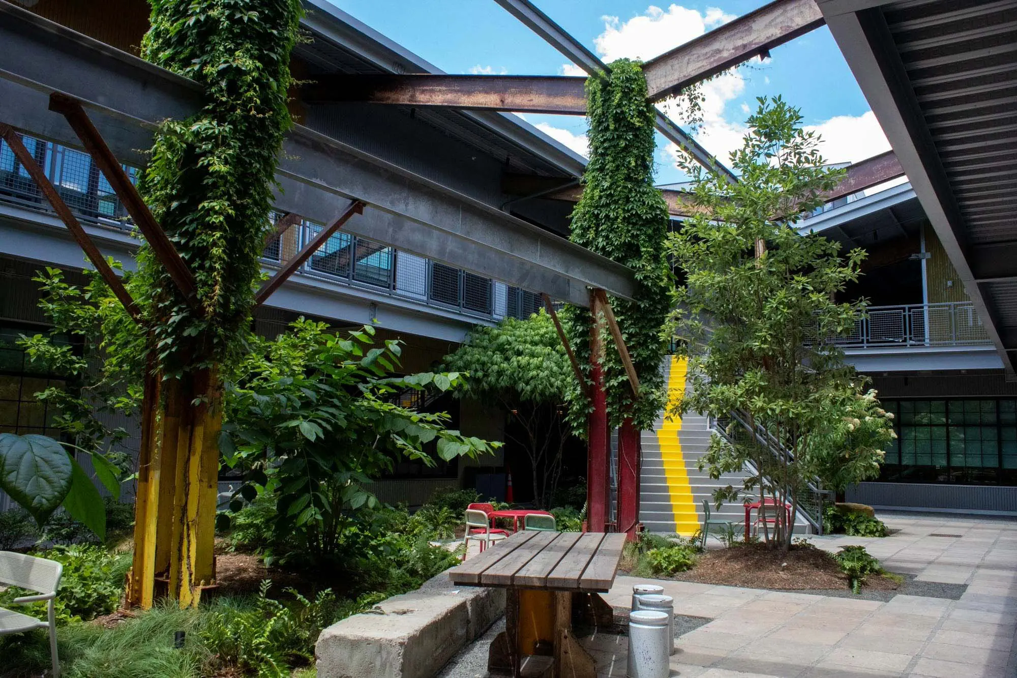 Outdoor courtyard with lush green plants, yellow painted beams, metal structures covered in vines, a wooden table, red and white chairs, and a staircase with a bright yellow stripe. Blue sky with clouds is visible above.