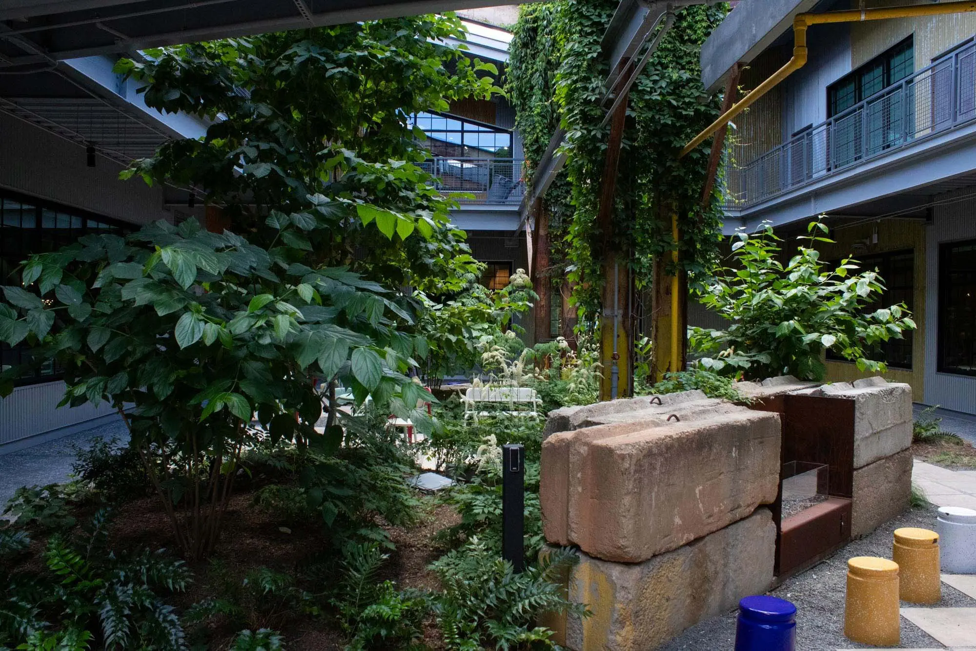 A lush courtyard garden with various green plants, trees, and ferns, surrounded by modern buildings and large windows. Stone blocks and colorful stools are in the foreground, with a balcony above.