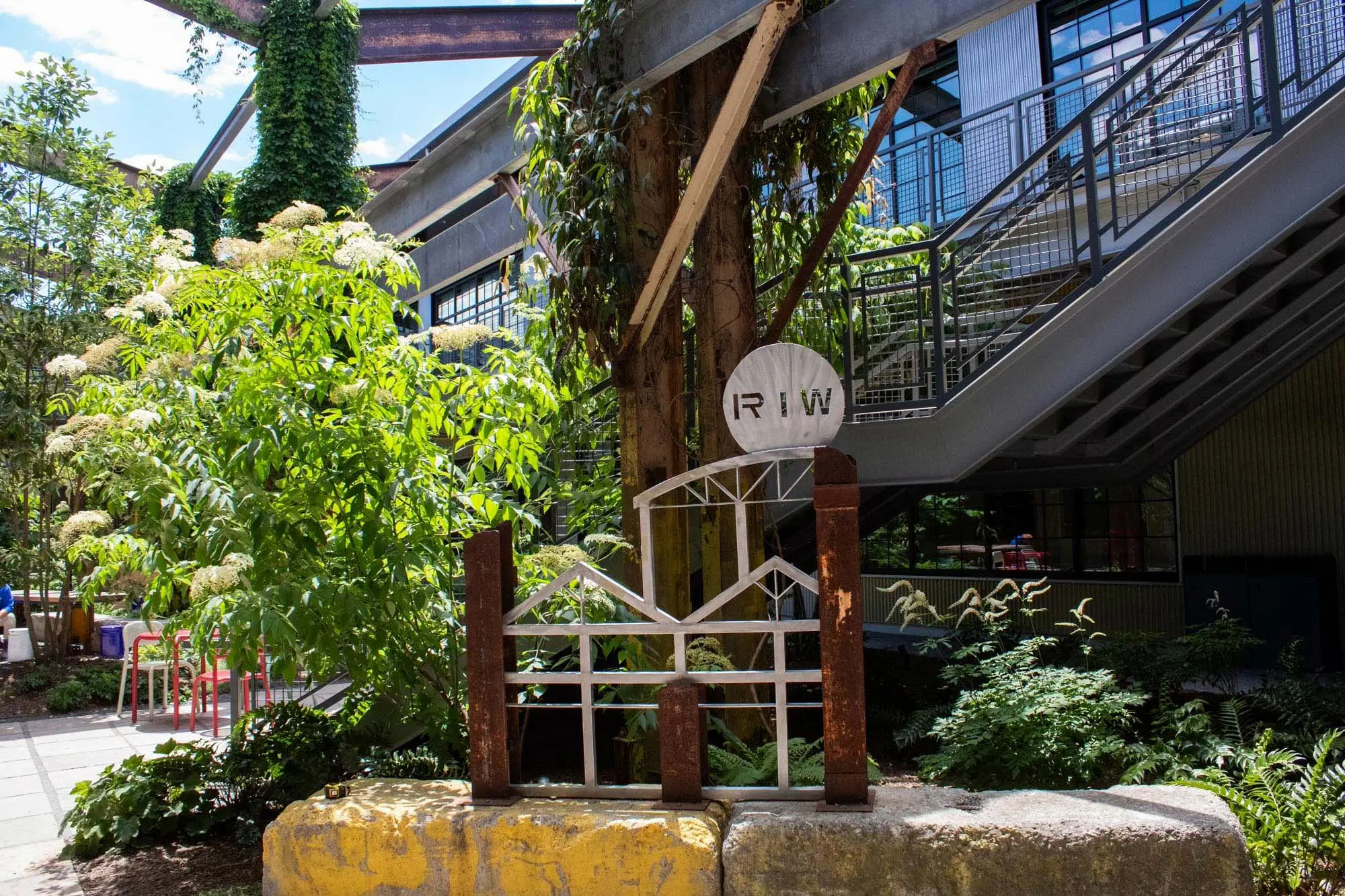 A metal sculpture labeled "RIW" stands by plants and trees in an outdoor area next to a building with large windows and a metal staircase. Sunlight filters through overhead beams and greenery.