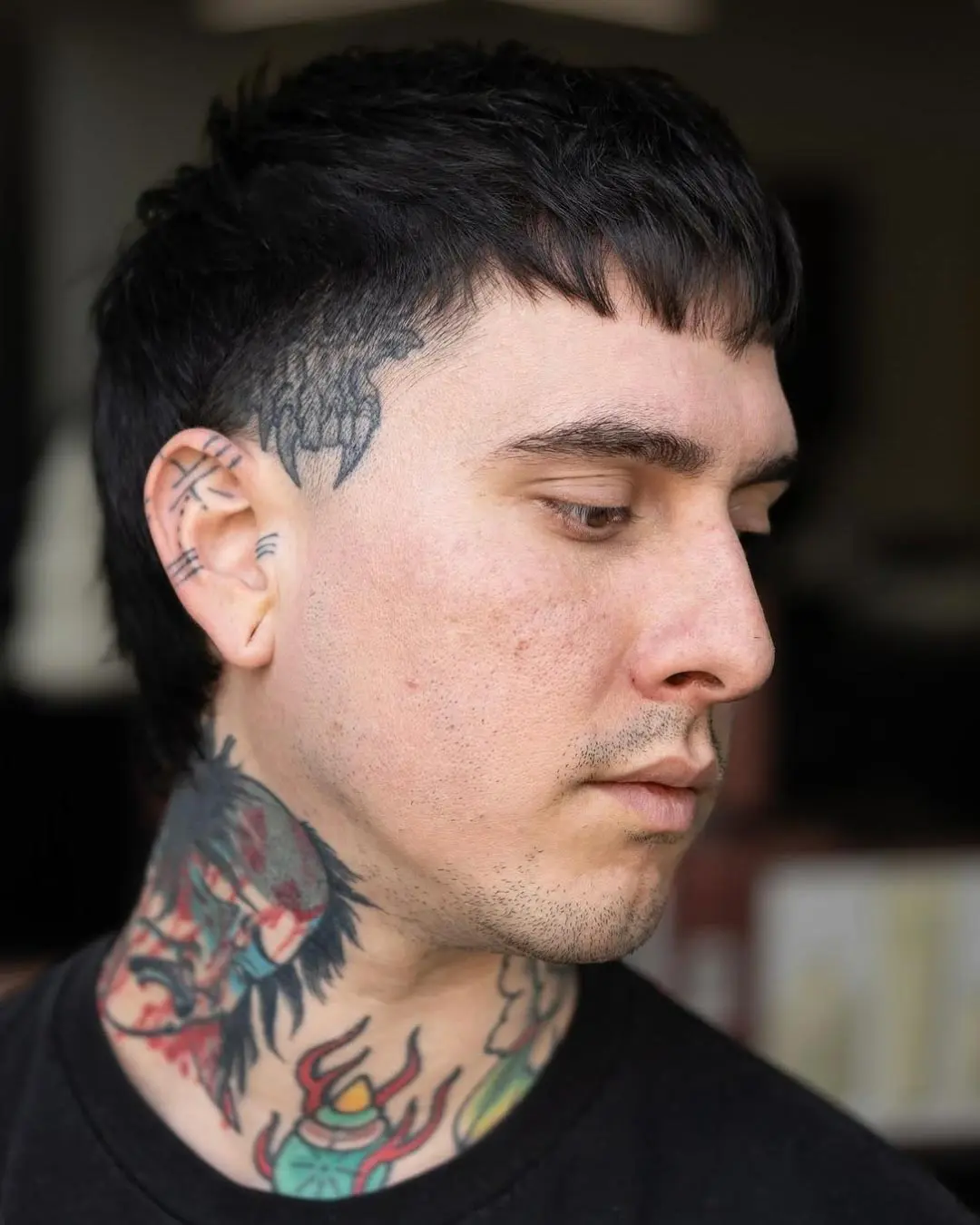 kyle-temple-portfolio-1. A person with short dark hair and tattoos on their neck and face looks down and to the side. The tattoos include designs behind the ear and a colorful figure on the neck. The background is blurred.