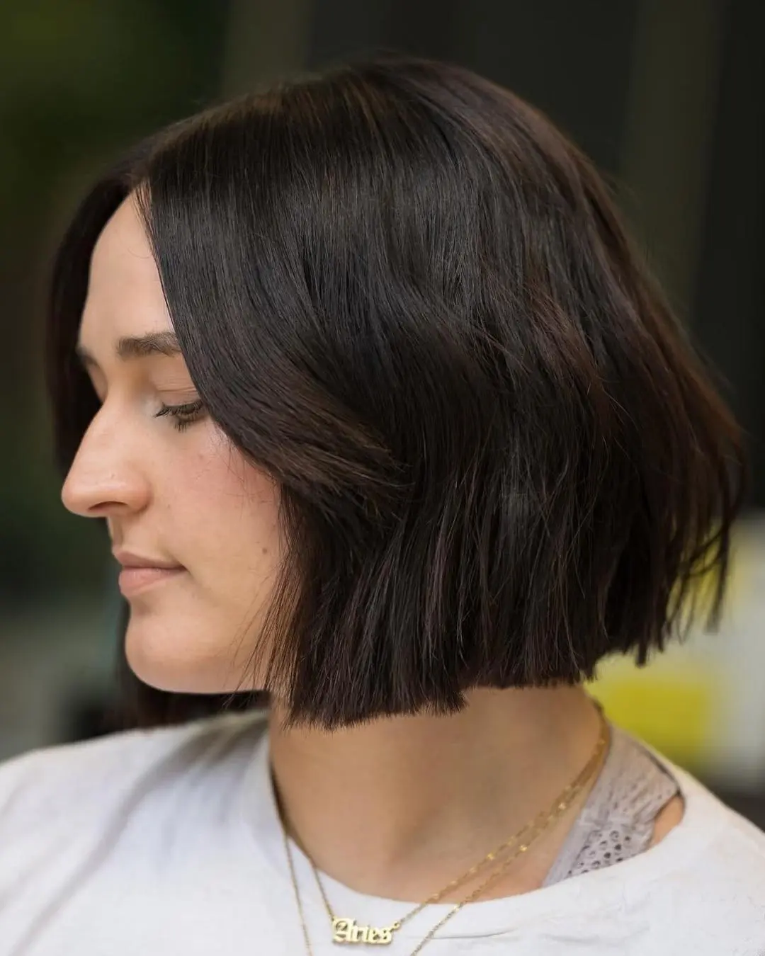 kyle-temple-portfolio-10 A person with straight, dark brown hair styled in a blunt, chin-length bob is shown in profile, wearing a white top and layered gold necklaces. The background is softly blurred.