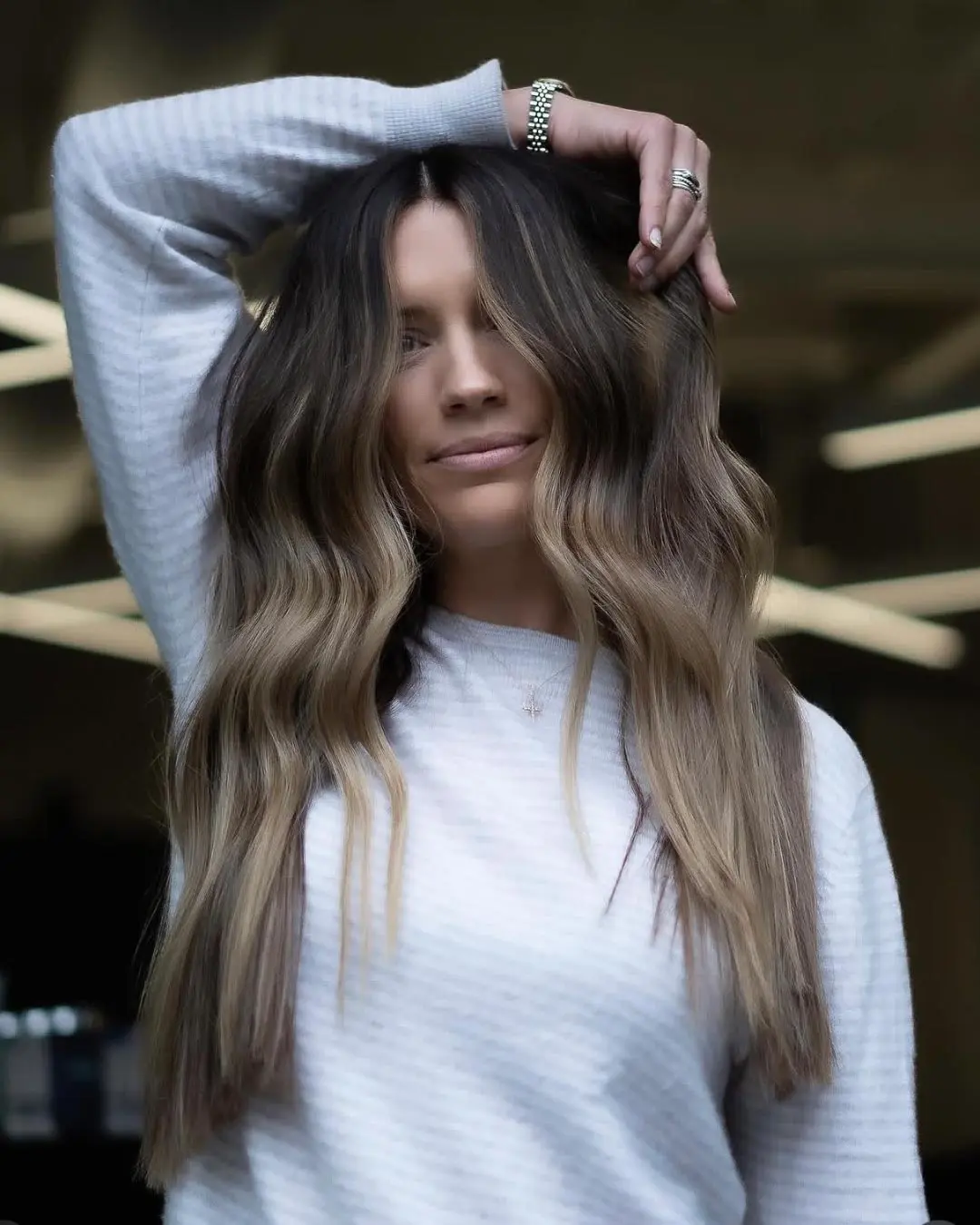 kyle-temple-portfolio-11 A woman with long, wavy, ombre hair wears a white textured sweater, posing with one arm raised and hand on her head, smiling slightly in a softly lit indoor setting.