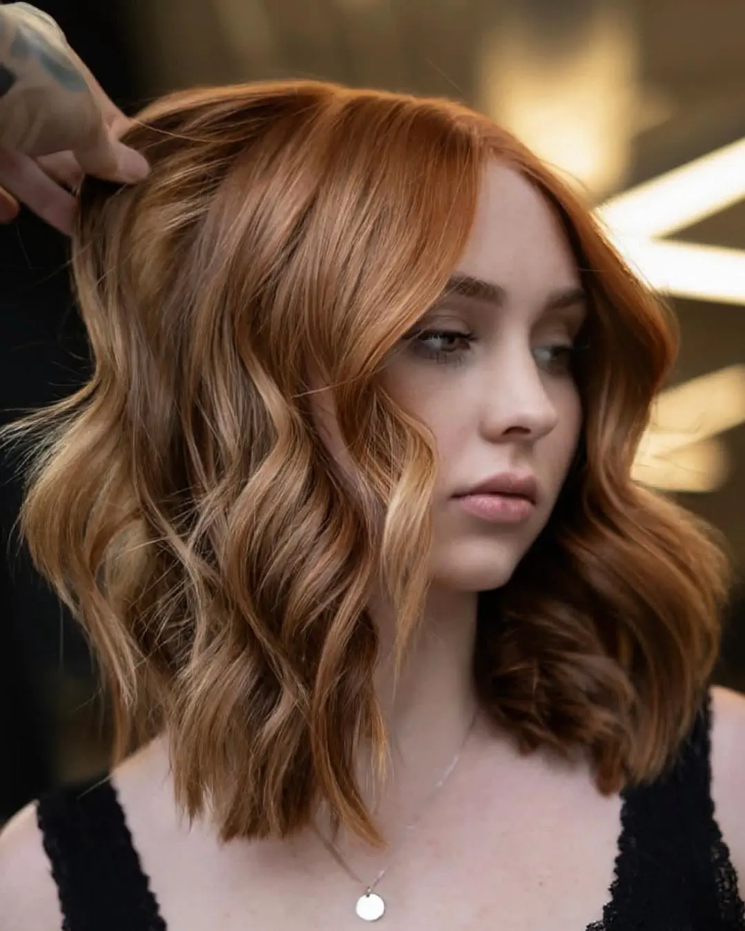 kyle-temple-portfolio-4 A woman with wavy, shoulder-length copper hair wears a black top and a silver necklace. Someone lifts a section of her hair to show its soft waves and color. The background is softly blurred with warm lighting.