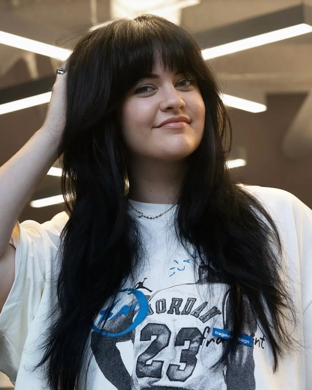 kyle-temple-portfolio-5 A young woman with long, dark hair and bangs smiles while holding her hair with one hand. She is wearing a white T-shirt with a "23" and "Jordan" graphic. Modern lighting fixtures are visible in the background.
