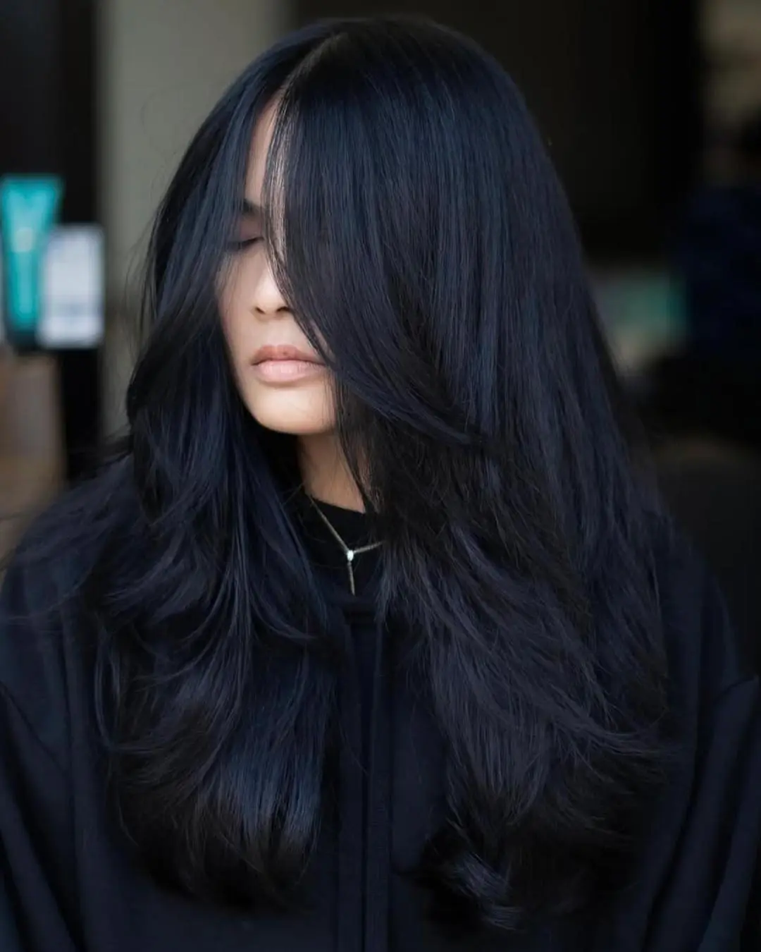 kyle-temple-portfolio-6 A woman with long, thick, straight black hair and curtain bangs wears a black top and looks down, partly obscured by her hair. The background is softly blurred.