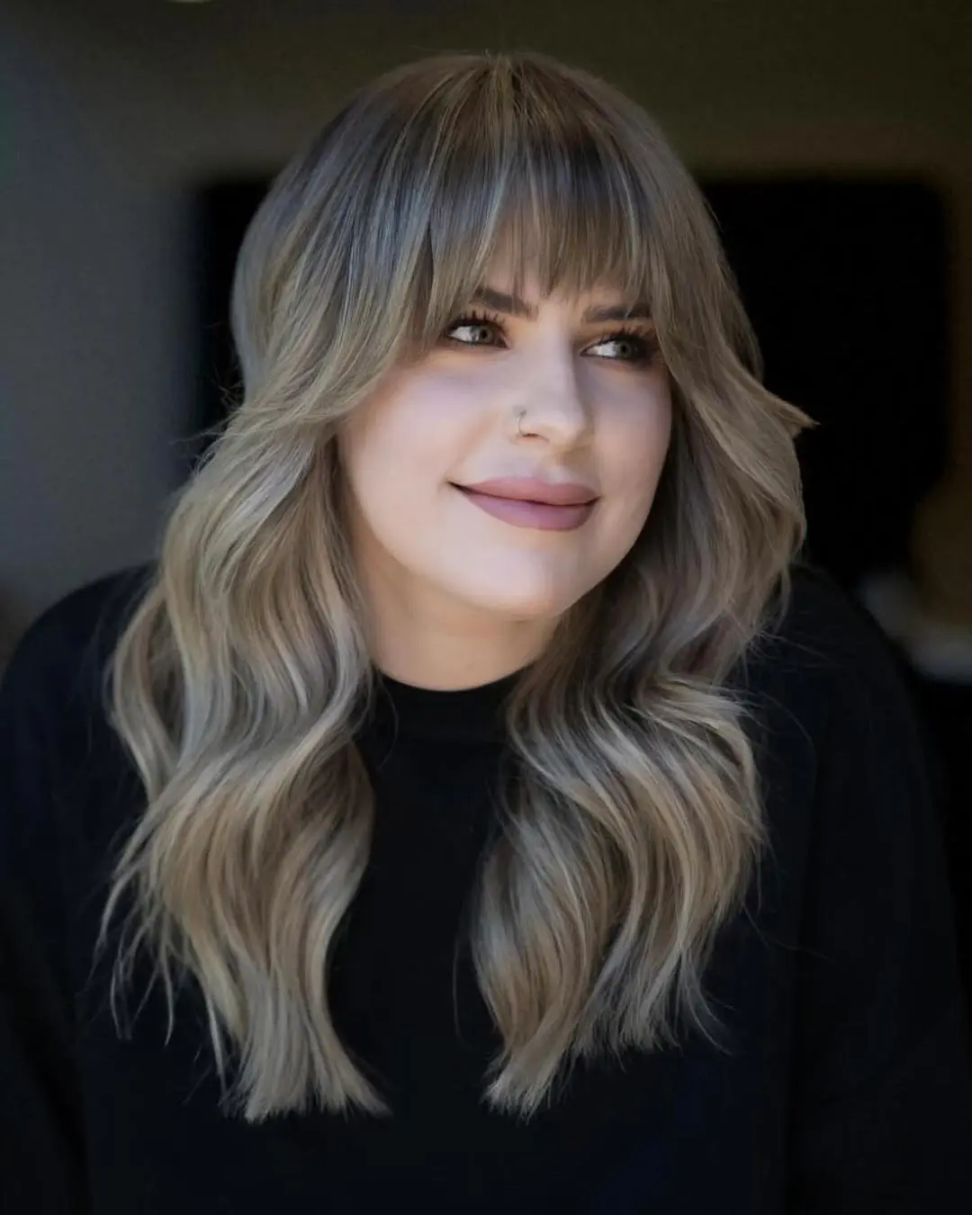 kyle-temple-portfolio-7 A woman with long, wavy, light brown hair with blonde highlights and bangs, wearing a black top, smiles softly while looking slightly to the side.