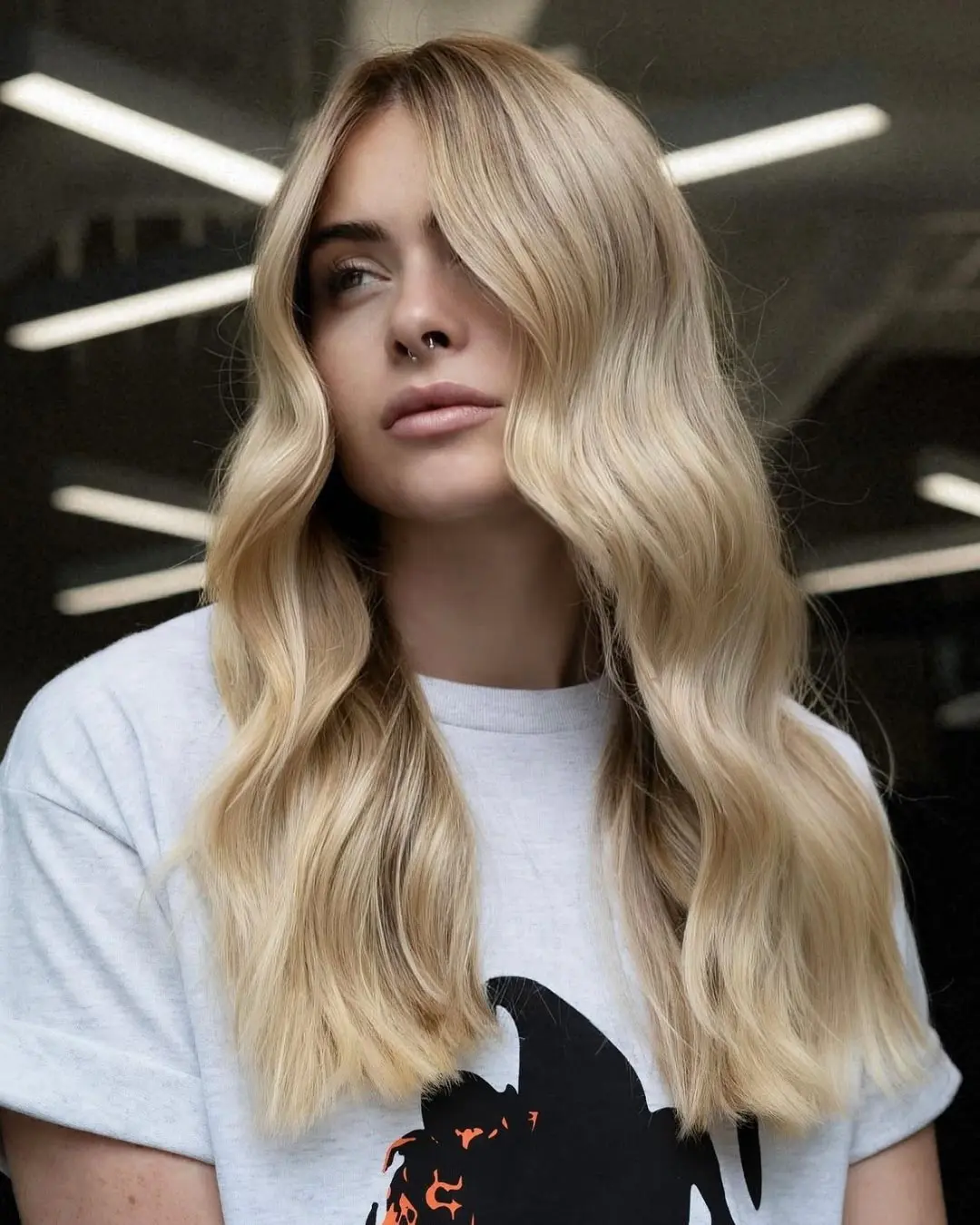 kyle-temple-portfolio-8 A woman with long, wavy, blonde hair wearing a white T-shirt stands indoors, facing slightly left. Bright lights and a blurred background are visible behind her.