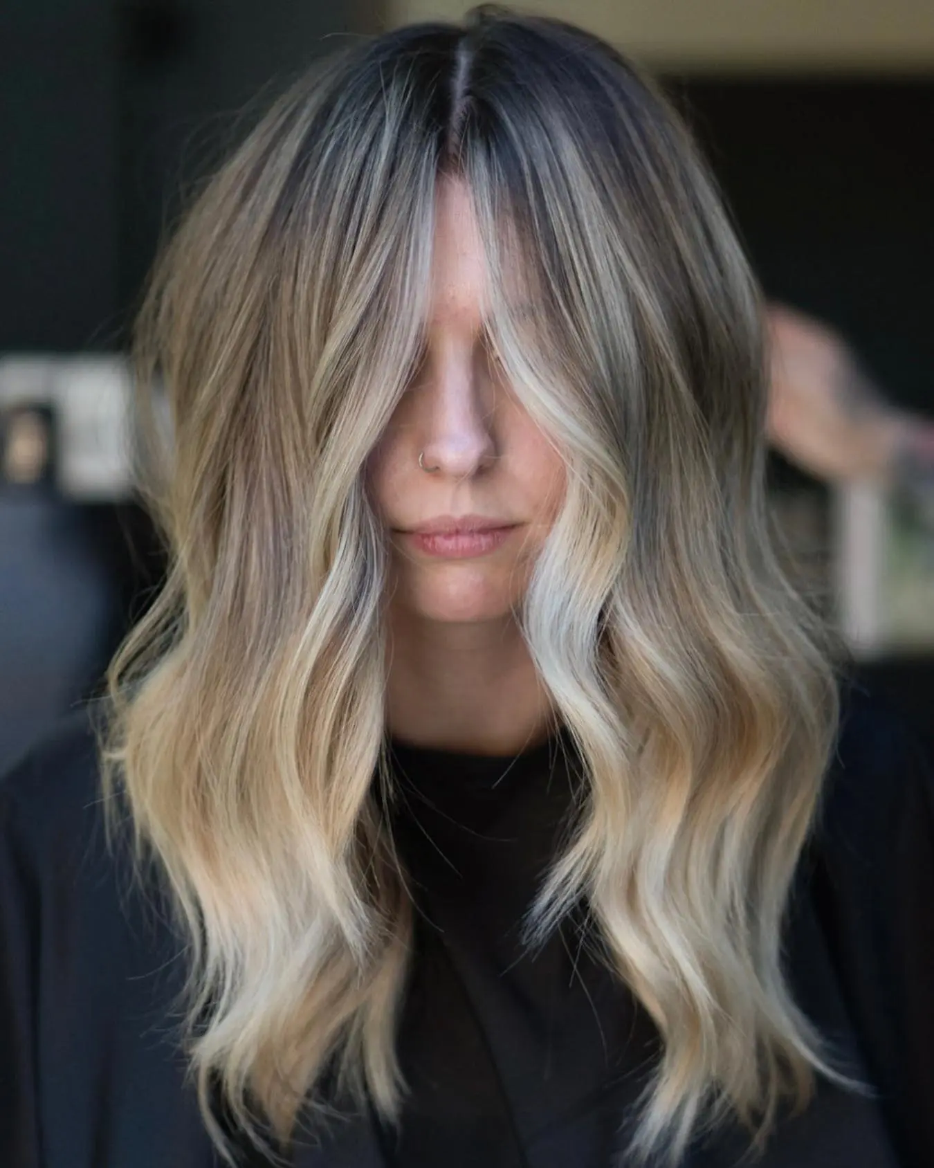 nathan-portfolio-1 A person with long, wavy, blonde hair featuring dark roots and lighter ends, parted in the middle with curtain bangs partially covering their face, wearing a black top.