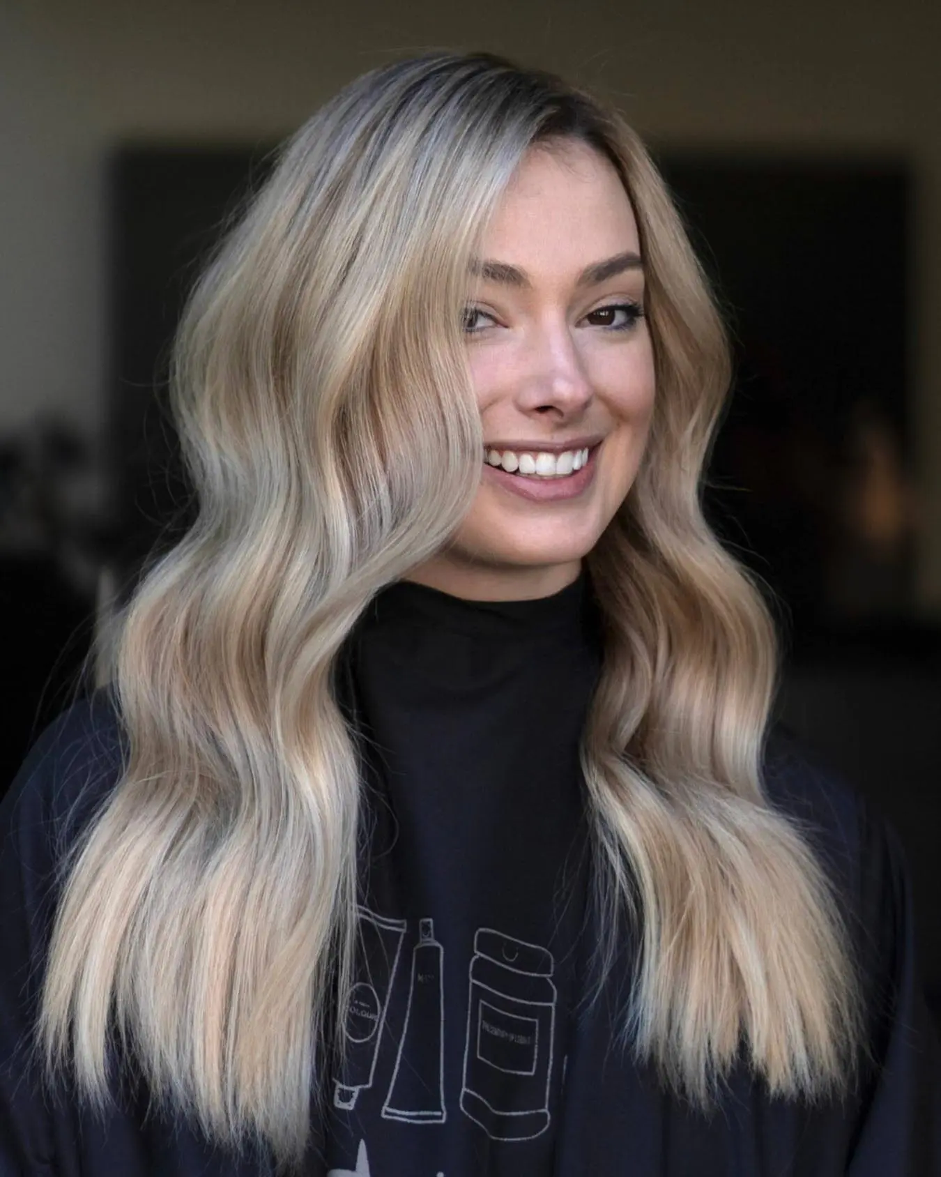 nathan-portfolio-2 A woman with long, wavy, blonde hair smiles at the camera. She is wearing a black salon cape, and her hair has soft, blended highlights. The background is softly blurred.
