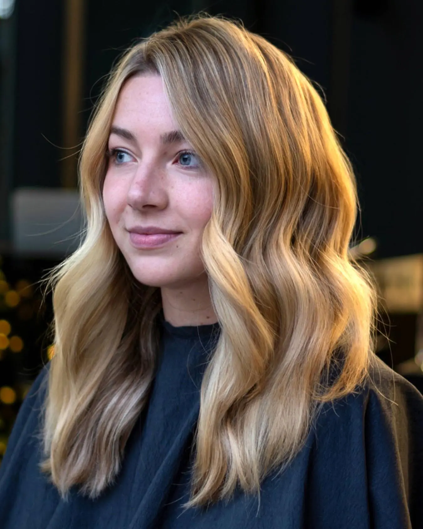 nathan-portfolio-3 A woman with long, wavy blonde hair and blue eyes looks slightly to the side, wearing a dark salon cape. The background is softly blurred, highlighting her freshly styled hair.
