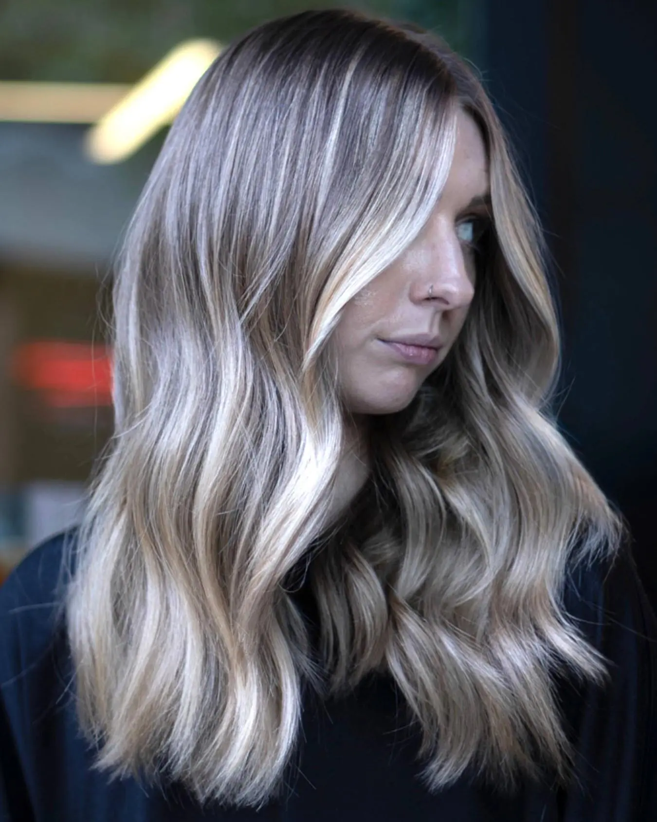 nathan-portfolio-4 A woman with long, wavy, ash-blonde hair featuring soft, blended highlights, parted slightly off-center, stands against a dark, blurred background.