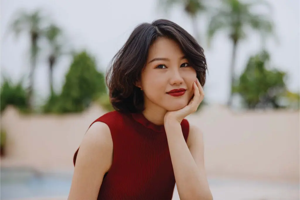 A woman with a fresh summer hair trim and short dark hair wears a sleeveless red top, resting her chin on her hand and smiling softly. She sits outdoors with blurred greenery and palm trees in the background.