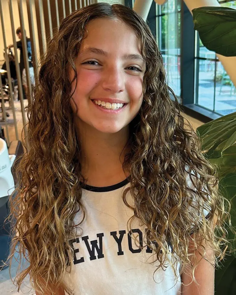 A long, naturally curly hair transformation created at Sunday Salon in Raleigh, featuring defined ringlets enhanced by warm golden blonde highlights and natural dimension.