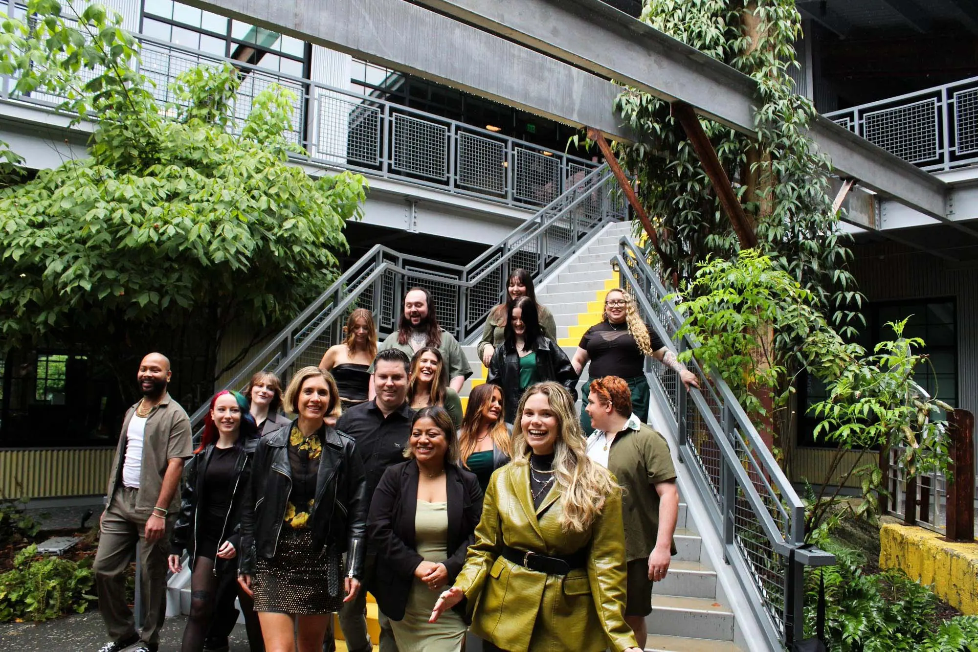 A group of people, smiling and walking down outdoor stairs, surrounded by greenery and industrial-style railings.