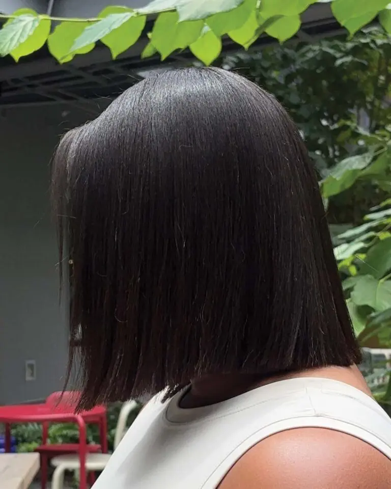 A person with straight, espresso dark brunette hair styled in a precision blunt bob stands outdoors, wearing a sleeveless white top. Sleek green leaves and some red and white chairs are visible in the background.