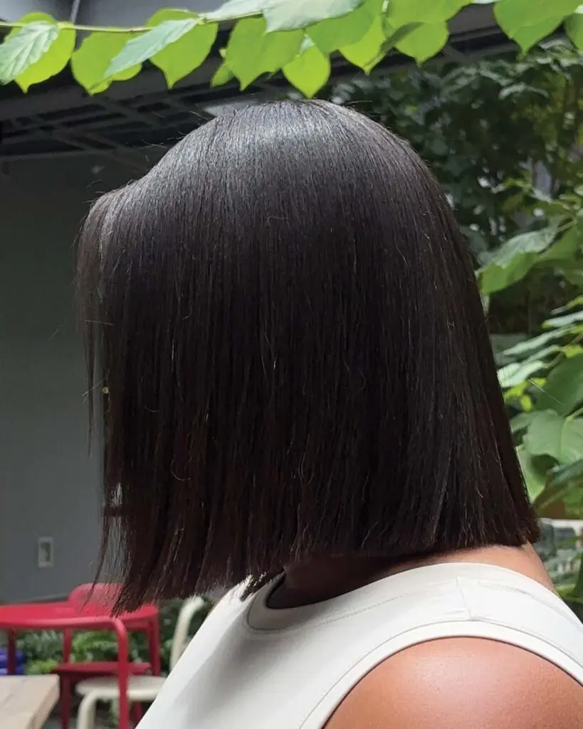 Sleek Espresso Dark Brunette Precision Blunt Bob A person with straight, espresso dark brunette hair styled in a precision blunt bob stands outdoors, wearing a sleeveless white top. Sleek green leaves and some red and white chairs are visible in the background.