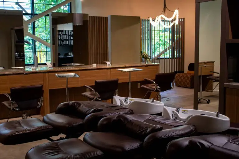 Modern hair salon interior with brown chairs, shampoo stations, large mirrors, and wood accents. Natural light streams in through large windows, and a chandelier hangs from the ceiling. The space appears tidy and inviting.