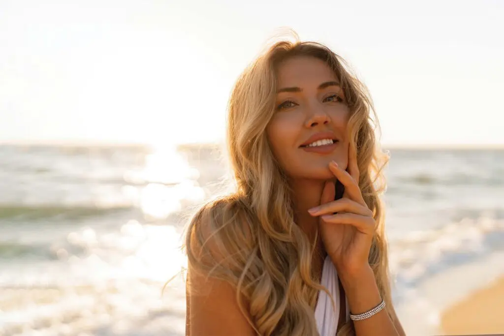 Young woman with long blonde hair styled in a low maintenance balayage smiles and touches her face while standing on a sunny beach, with ocean waves and bright sunlight in the background.