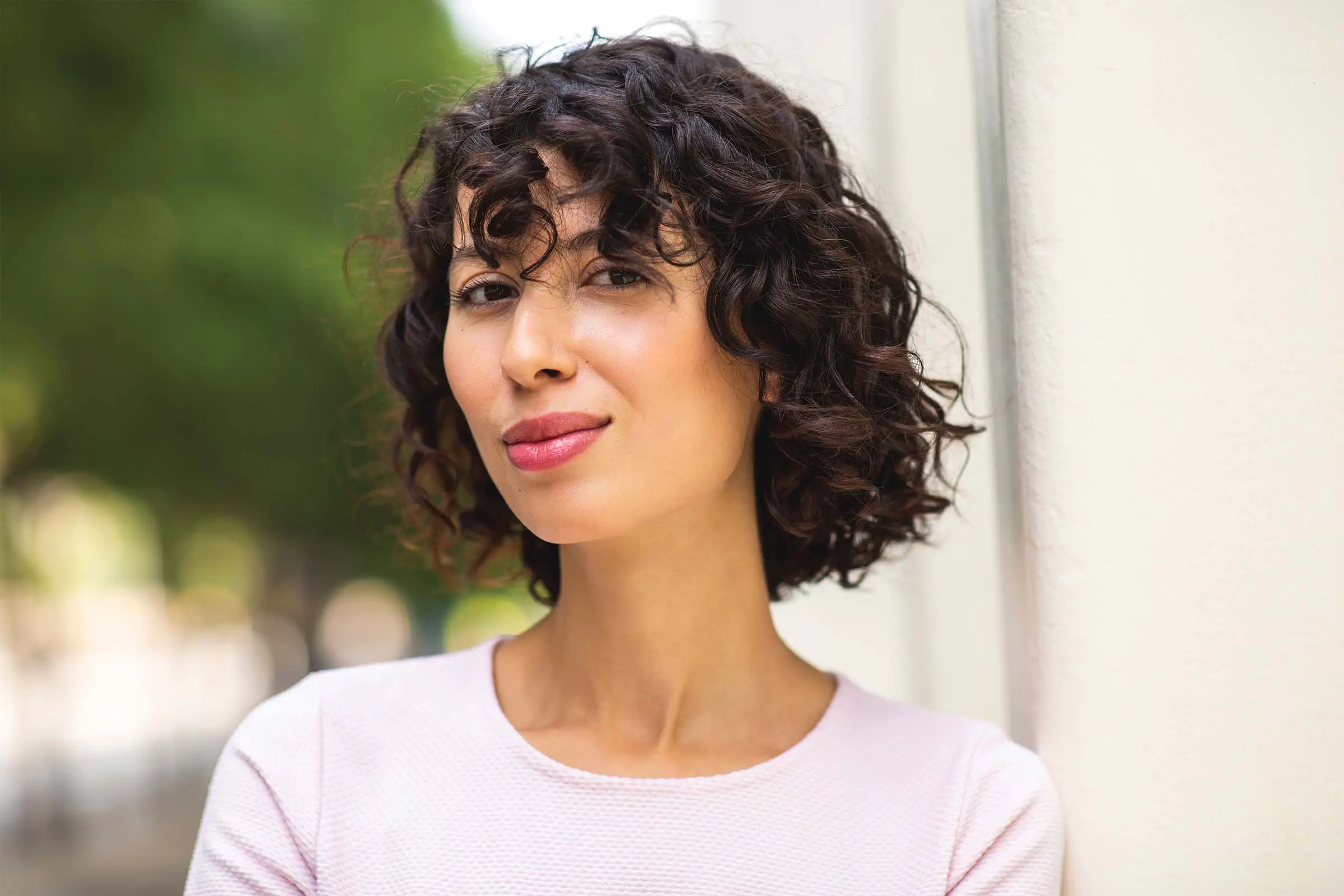 A portrait of a woman with chin-length, dark curly hair and bangs, smiling gently at the camera. Her curly hair frames her face as she wears a light pink shirt and stands against a white wall outdoors, with a soft-focus background of trees.