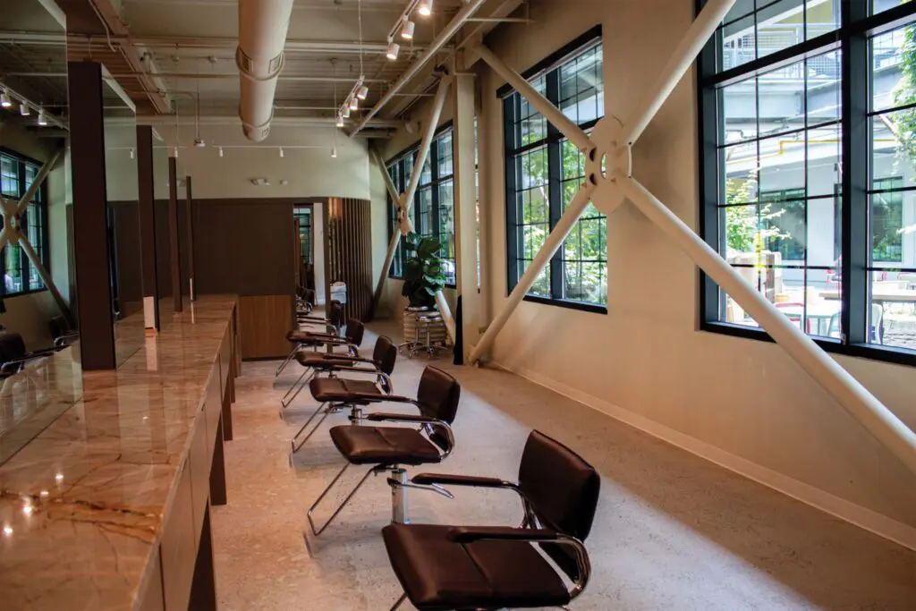 A modern, well-lit hair salon offering quality salon services, with empty brown chairs lined up along a marble countertop, large windows, exposed beams, and greenery visible outside.