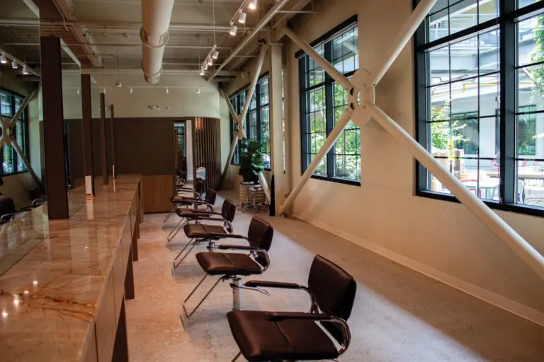 A modern, well-lit hair salon offering quality salon services, with empty brown chairs lined up along a marble countertop, large windows, exposed beams, and greenery visible outside.