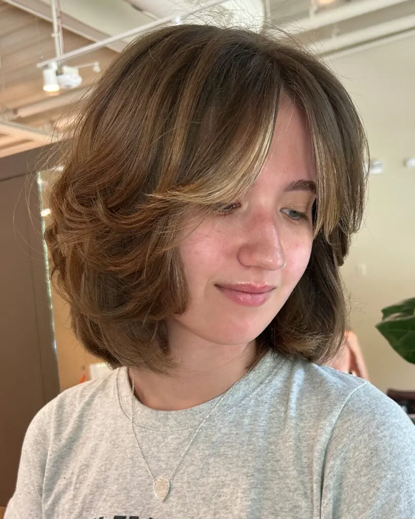 A woman displaying a chic, textured medium bob with soft curtain bangs and golden highlights styled by Sunday Salon in Raleigh.